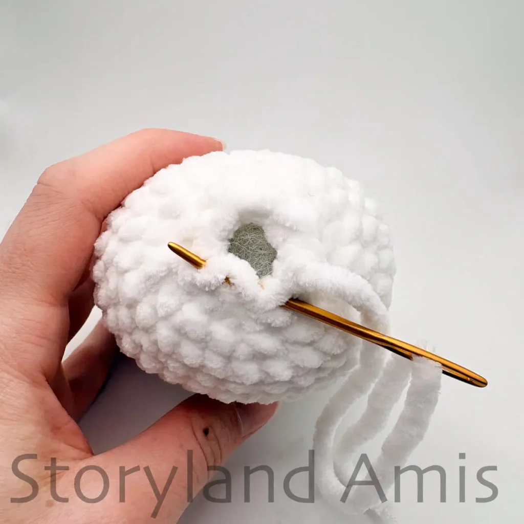 A close-up of a gold tapestry needle being used to sew a hole closed on the top of an amigurumi ghost toy.