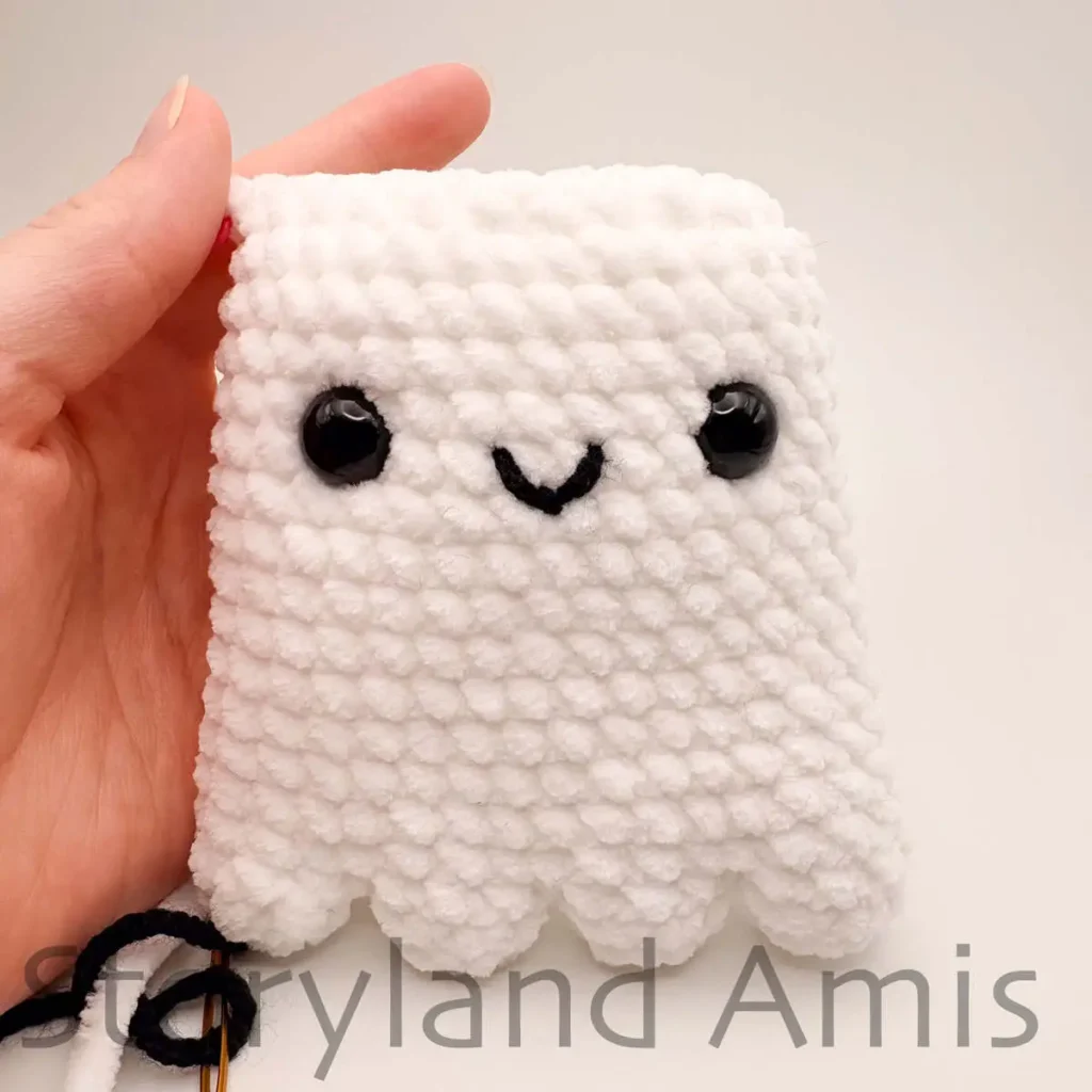 A hand holds a partially finished amigurumi ghost toy with two black plastic safety eyes attached to its front and a black embroidered mouth.