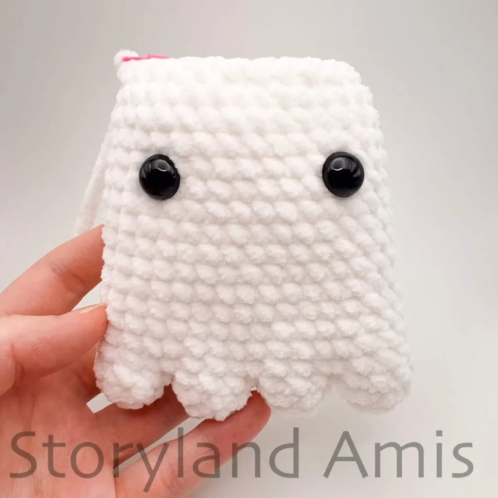 A hand holds a partially finished amigurumi ghost toy with two black plastic safety eyes attached to its front.