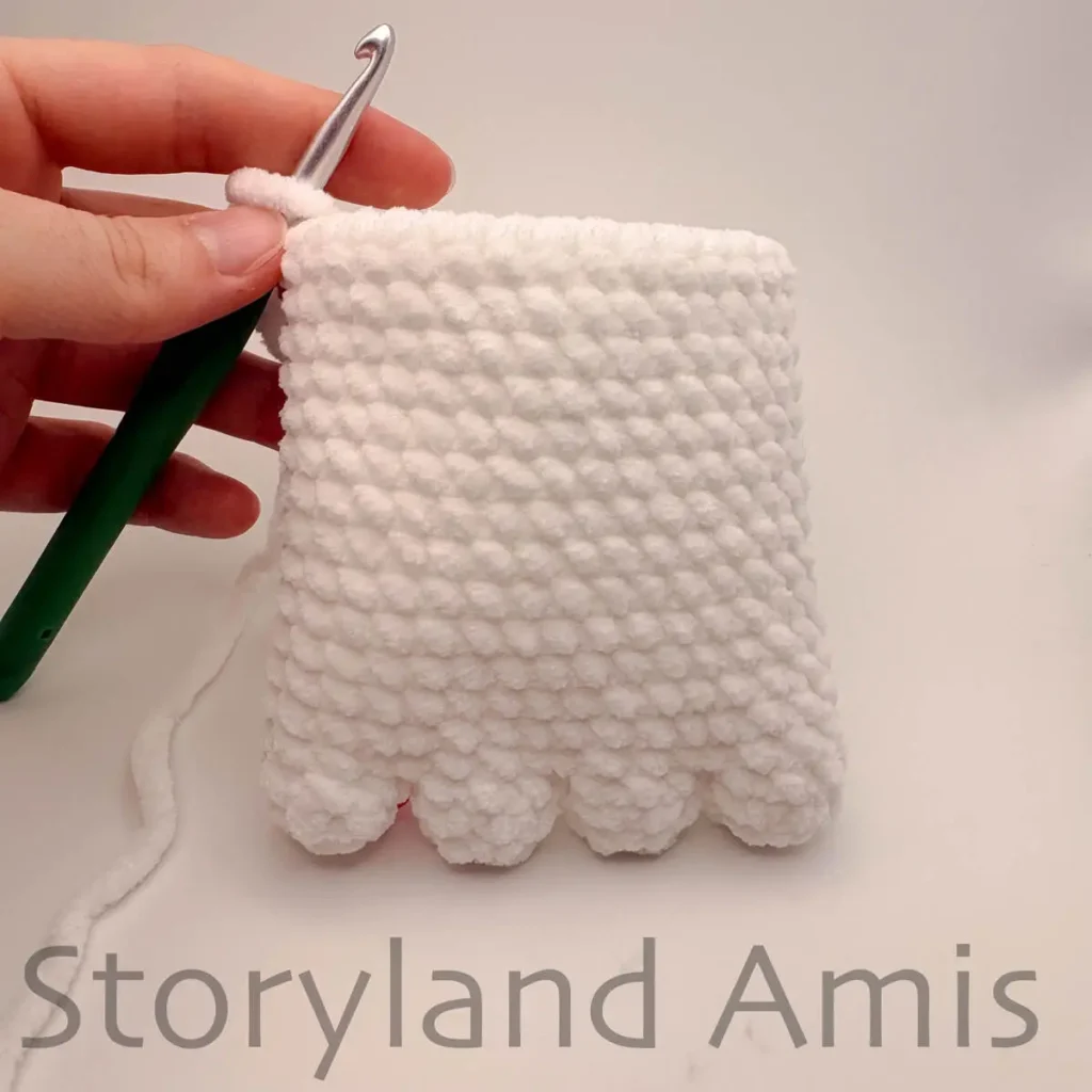 A hand holds a partially crocheted ghost body steady with a green crochet hook attached to the corner stitch of the body.