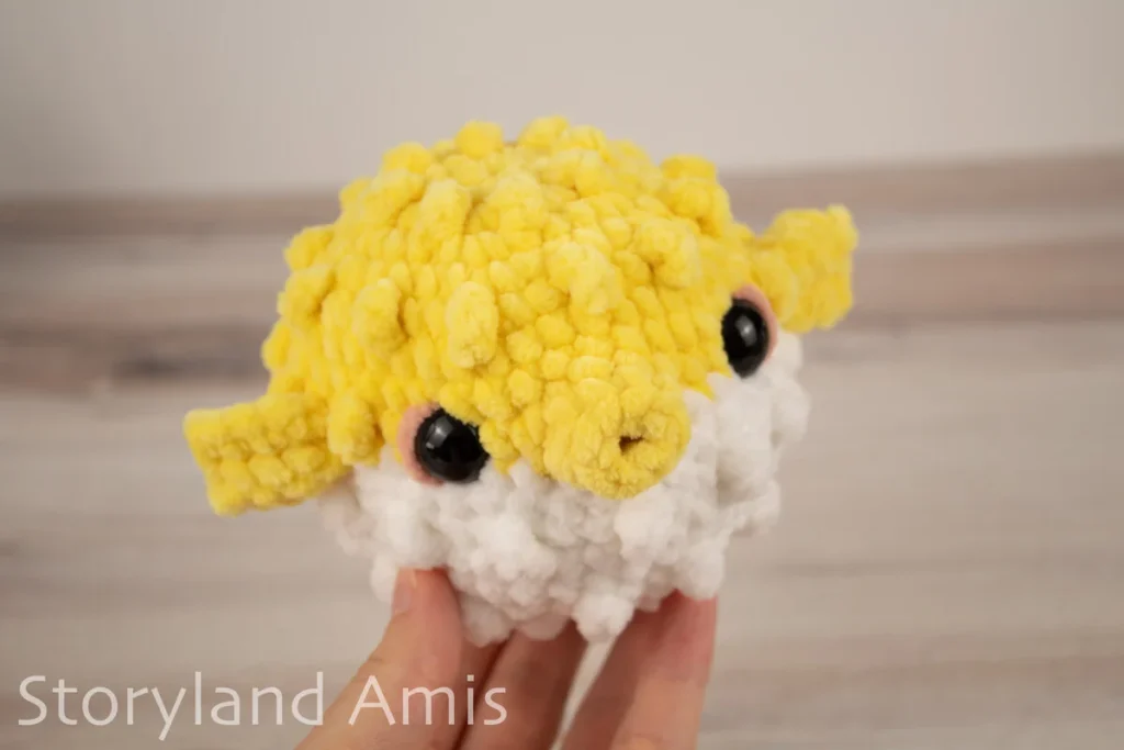 A hand holds an amigurumi pufferfish toy against a wooden background.