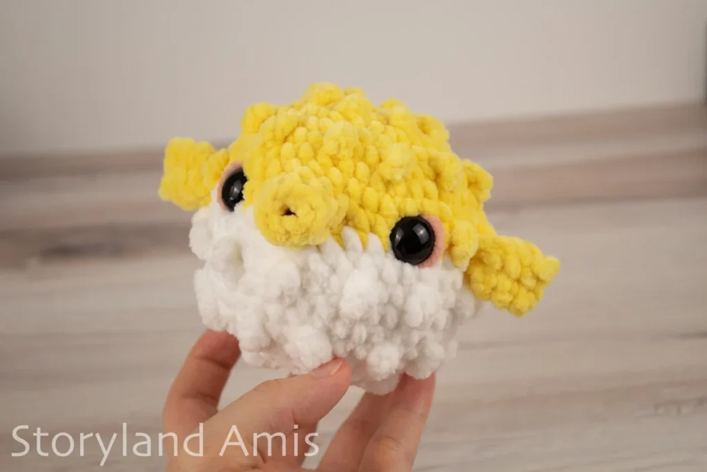 A hand holds an amigurumi pufferfish toy against a wooden background.