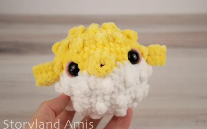 A hand holds an amigurumi pufferfish toy against a wooden background.